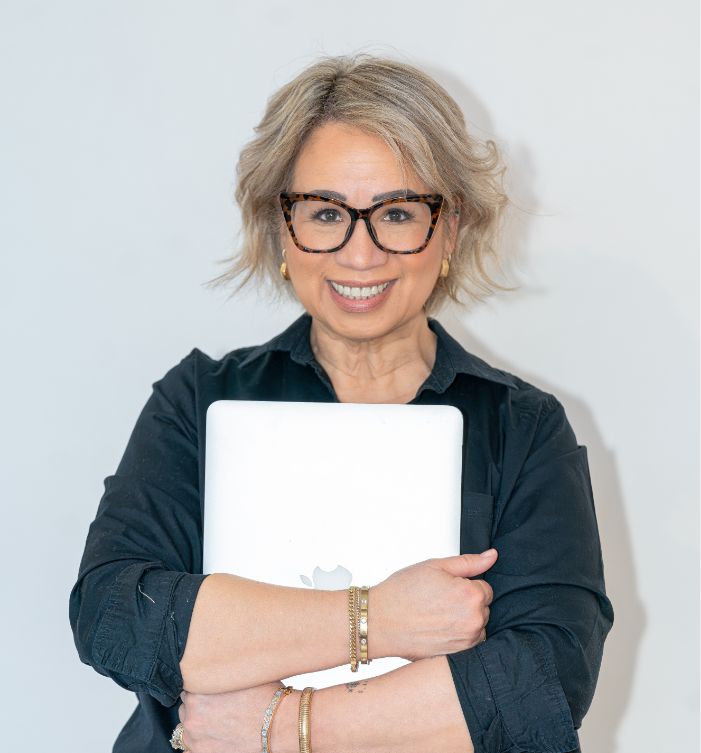 Smiling woman with short, blonde hair and glasses, wearing a black shirt, holding a closed silver laptop with both arms against a plain light background.