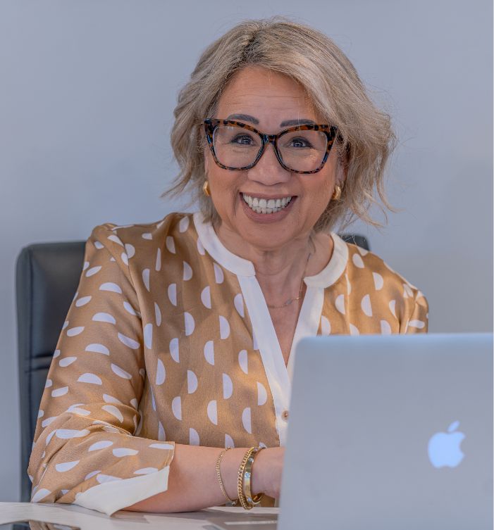 Smiling older woman with short, light hair and glasses sits at a desk in front of an open laptop. She wears a brown and white patterned blouse and gold bracelets, with a neutral background.