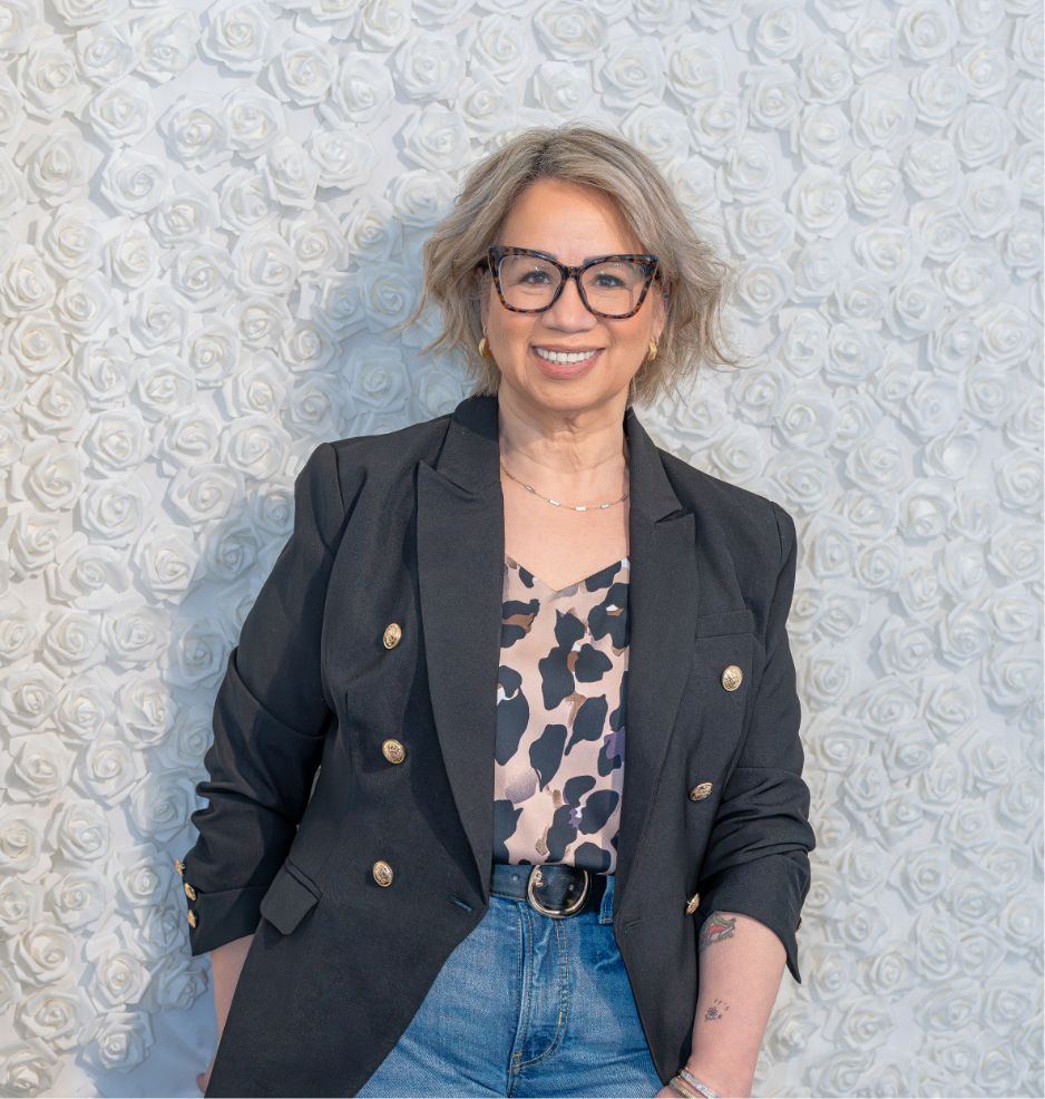 A smiling person with short, grayish hair and glasses wears a black blazer, animal print top, and blue jeans. They stand in front of a textured white wall with a raised rose pattern.