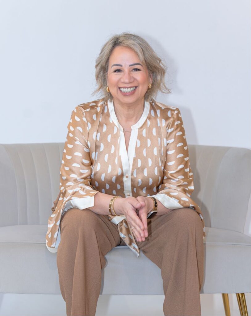 A smiling woman with short gray hair sits on a light gray sofa. She wears a brown and white patterned blouse and brown pants, with her hands clasped loosely in front of her. The background is plain and light-colored.