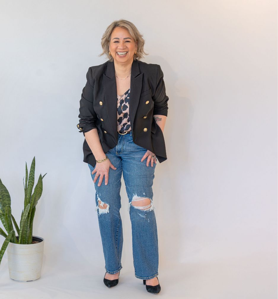A woman with short hair smiles while standing against a plain background. She wears a black blazer, a leopard print top, ripped blue jeans, and black heels. A potted plant sits on the floor to her left.